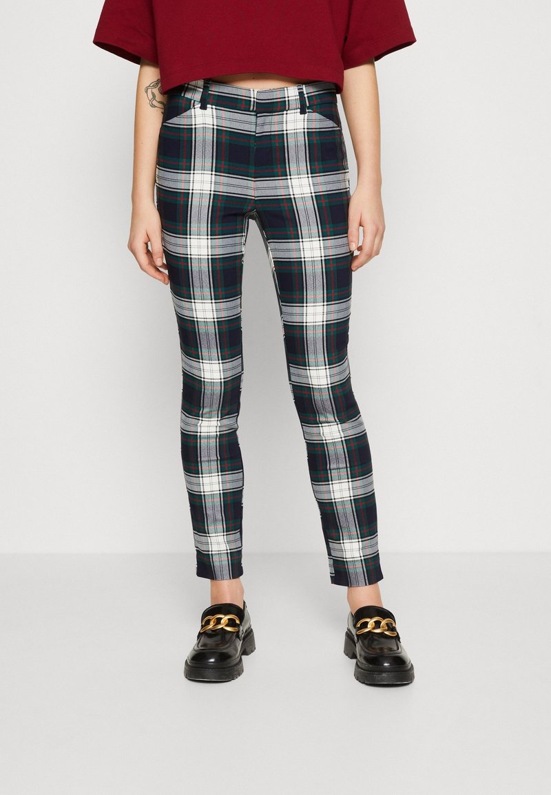 Personne portant un pantalon à carreaux noirs, blancs et verts, des mocassins noirs avec des chaînes dorées, et un haut rouge court, debout devant un fond uni.