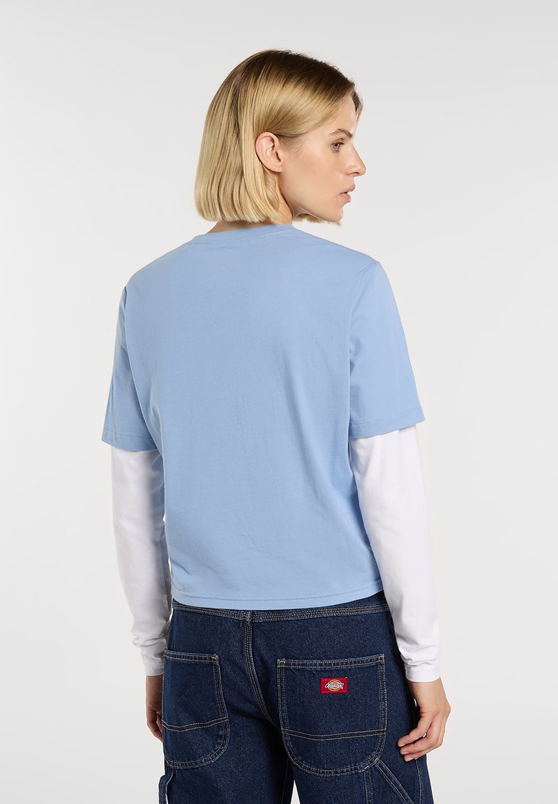 Light blue short-sleeve t-shirt, cropped design with a round neckline. Worn over a white long-sleeve shirt. Denim with patch pockets visible.