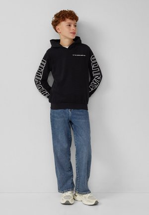 Ragazzo con capelli ricci che indossa un hoodie nero con "ULTIMATE" sulle maniche, jeans blu e sneakers bianche, in piedi contro un muro grigio chiaro.