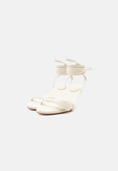 Ζευγάρι λευκών φλατ σανδαλιών με λεπτά λουράκια στα δάχτυλα και λουράκια στον αστράγαλο που τυλίγονται και δένουν γύρω από τον αστράγαλο, παρουσιάζονται σε ομοιόμορφο φόντο.