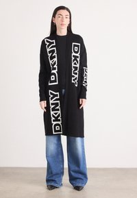 Črn dolg kardigan z belim tekstom "DKNY" na rokavih in spredaj, nosi se preko črne majice in je kombiniran s svetlo modrimi širokimi hlačami.
