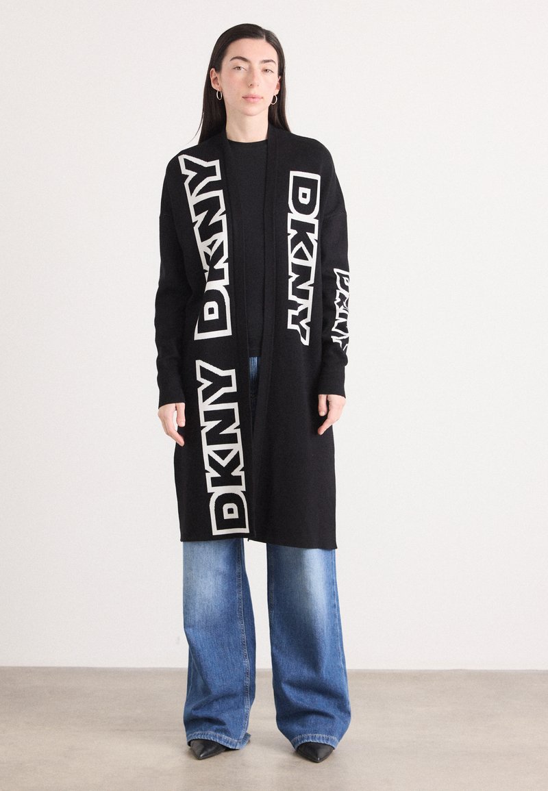 Črn dolg kardigan z belim tekstom "DKNY" na rokavih in spredaj, nosi se preko črne majice in je kombiniran s svetlo modrimi širokimi hlačami.