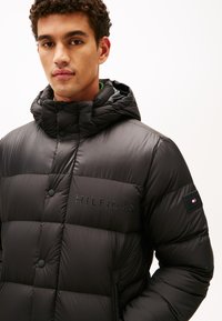 Sort puffer-jakke med hætte, der har vandrette quiltede sektioner, knapper og et prægede logo. Sidelommer med lynlås og struktureret stof.