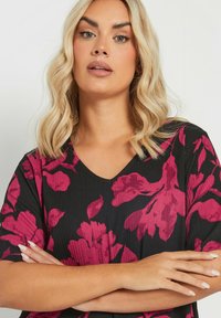 Top de tela negra con escote en V, que presenta grandes patrones florales en rosa y una superficie texturizada. La modelo cruza los brazos, mostrando los detalles del diseño.