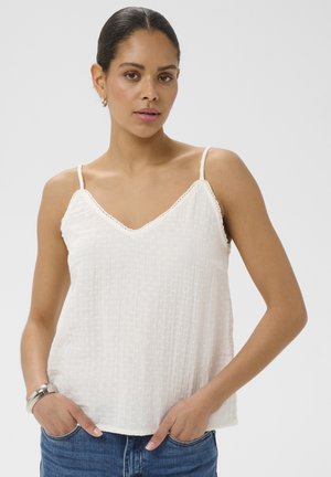 Femme aux cheveux foncés portant un haut sans manches blanc texturé et un jean bleu, debout les mains dans les poches devant un fond uni.