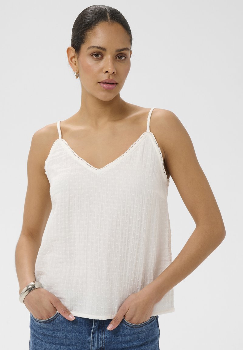 Femme aux cheveux foncés portant un haut sans manches blanc texturé et un jean bleu, debout les mains dans les poches devant un fond uni.
