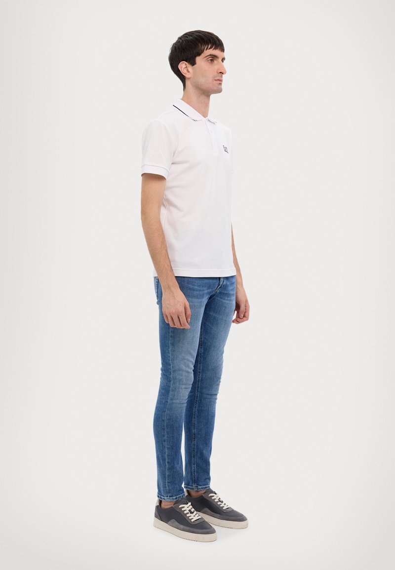 White polo shirt with short sleeves, navy accents on the collar, paired with light blue skinny jeans and grey sneakers.