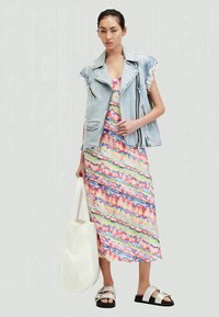 Dżinsowa kamizelka z postrzępionymi brzegami na kolorowej sukience maxi w tie-dye. Sukienka ma żywe paski w kolorach różowym, niebieskim i zielonym. Biała torba tote.