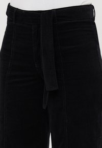 Black corduroy pants with a tie belt feature, high waist, vertical rib texture, and side pockets. Zipper closure at the waist.