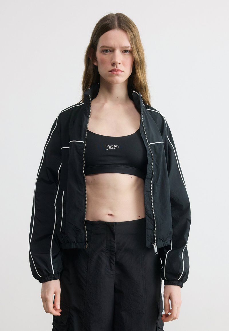 Jeune femme debout devant un fond blanc, portant une veste noire zippée avec des bordures blanches, un crop top noir et un pantalon noir.