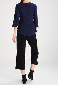 Top blu navy con scollo rotondo e maniche a campana 3/4, con dettaglio a occhio di bottone sul retro. Abbinato a pantaloni neri a tre quarti.