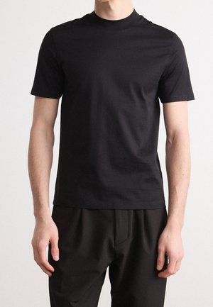 Man draagt een effen zwarte T-shirt met korte mouwen en een ronde hals en een zwarte broek, met ontspannen armen langs zijn zij.