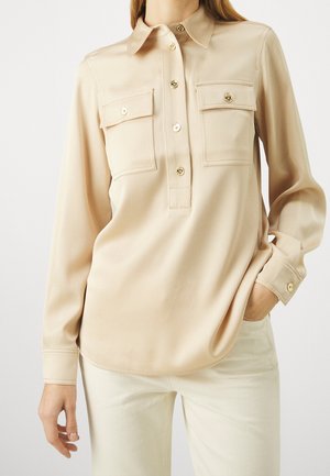 Beige Bluse mit langen Ärmeln, vorderer Knopfleiste und zwei Brusttaschen, kombiniert mit hellen Hosen.