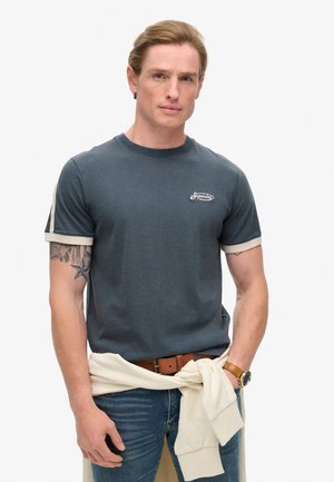 Navy blue short-sleeve t-shirt with cream accents and a logo on the chest, paired with light jeans and a cream sweater tied around the waist.