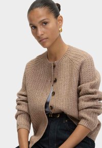 Mujer con el cabello hacia atrás, llevando pendientes de aro dorados, un cárdigan de punto beige abotonado, una blusa blanca y jeans oscuros, frente a un fondo liso.