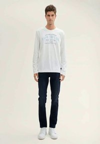 Camiseta de algodão branca de manga longa com texto gráfico azul, combinada com jeans escuros e sapatilhas brancas, de pé contra um fundo simples.