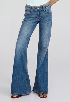 Vrouw draagt blauwe jeans met hoge taille en uitlopende pijpen, gecombineerd met rode sandalen met open teen op een effen grijze achtergrond.