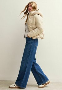 Beige pufferjack met een capuchon, brede blauwe denim jeans en witte sneakers met bruine accenten. Zachte texturen en een casual ontwerp.