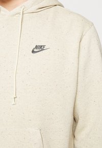 Béžová mikina s kapsou vpředu, kapucí se stahovací šňůrkou a černým logem Nike na levé hrudi.