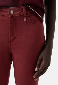Burgundy fitted jeans with a smooth texture, featuring a zip fly, button closure, and slight stretch for comfort.