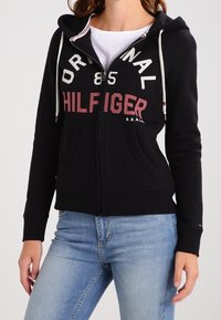 Svart zip-up hoodie med vit och röd "HILFIGER"-text över bröstet, med dragsko i huvan och framfickor. Material av bomullsblandning.