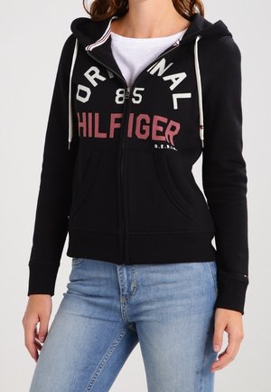 Felpa nera con zip, con la scritta "HILFIGER" in bianco e rosso sul petto, dotata di cappuccio con cordini e tasche frontali. Materiale misto cotone.