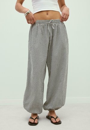 Osoba nosząca luźne czarno-białe spodnie w kratę z elastycznymi mankietami, biały krótki top, bransoletki i czarne japonki, stojąca w pomieszczeniu.