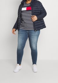 Person som bär en marinblå pufferjacka över en randig skjorta i marinblått, vitt och rött, tillsammans med blå skinny jeans och vita sneakers.