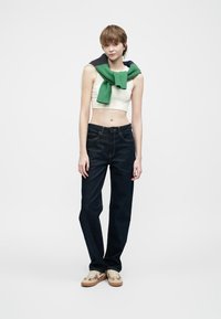 Kratka bela majica na bretele, temno modri traper hlače ravnega kroja, zeleni pleteni pulover čez ramena in svetle športne copate.