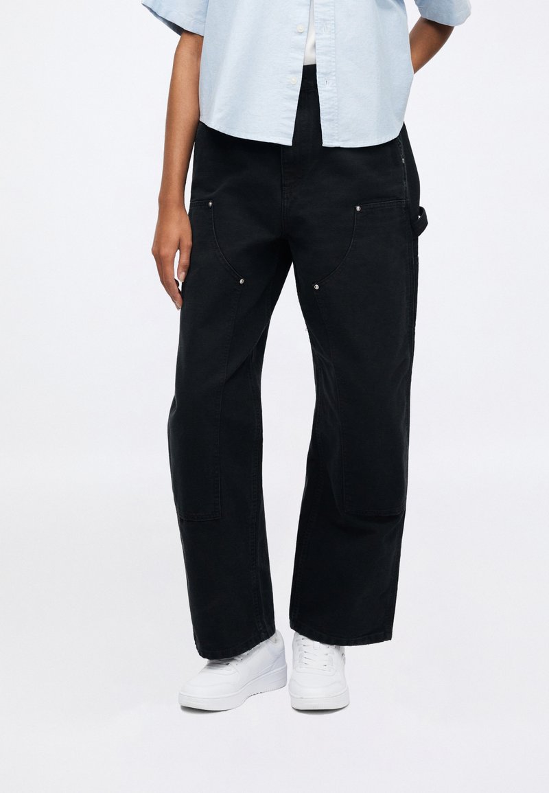 Pantalon noir surdimensionné avec une coupe décontractée, doté de poches avant, coutures visibles et accents de quincaillerie argent ée. Associé à des baskets blanches.