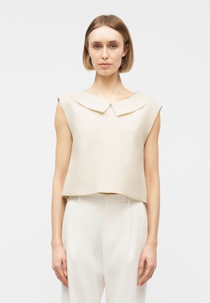 Femme aux cheveux blonds courts portant un haut beige sans manches à col large et un pantalon blanc taille haute, debout devant un fond uni.