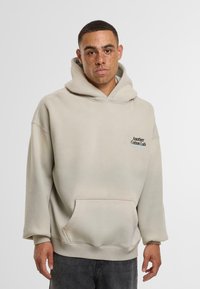 Giovane con lentiggini che indossa un hoodie beige chiaro con il testo "Another Cotton Lab" sul petto e jeans scuri, in piedi davanti a uno sfondo uniforme.