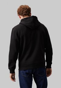 Felpa nera con un ampio cappuccio, polsini e vita a coste, realizzata in un morbido tessuto; indossata sopra un paio di jeans in denim blu, vista da dietro.