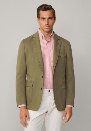 Olivgrüner Blazer mit Reverskragen, zwei Vordertaschen und schwarzen Knöpfen. Getragen über einem hellrosa und weiß gestreiften Hemd und weißen Hosen.
