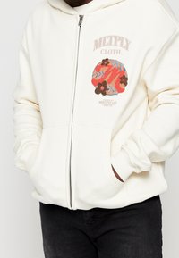 Sudadera con capucha blanca con cremallera, que presenta un diseño colorido de flores y figuras abstractas en el pecho, con puños acanalados y capucha forrada.