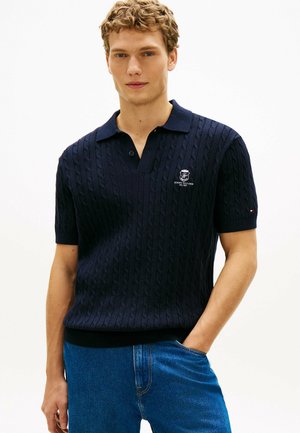 Junger Mann, der ein marineblaues Polohemd mit Zopfmuster und gesticktem Logo sowie blaue Jeans trägt, steht mit einer Hand in der Tasche.