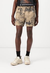 Beige Shorts mit einem dunklen botanischen Muster, elastischem Bund, seitlichen Taschen und einem kleinen Logo auf der Vorderseite. Kombiniert mit hellen Sneakers und schwarzen Socken.