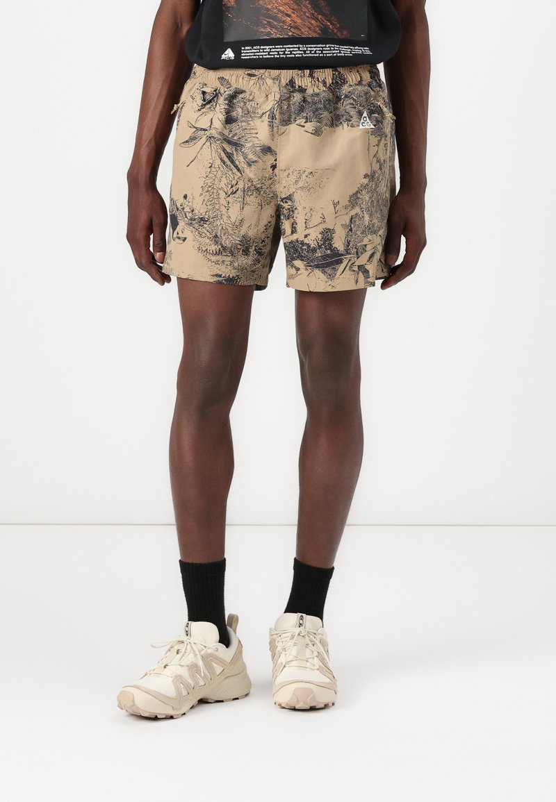 Beige Shorts mit einem dunklen botanischen Muster, elastischem Bund, seitlichen Taschen und einem kleinen Logo auf der Vorderseite. Kombiniert mit hellen Sneakers und schwarzen Socken.