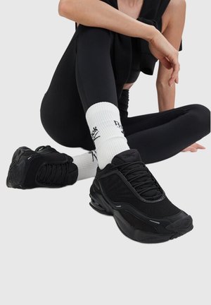Person iført sorte sportssko, hvide Reebok-sokker og sorte leggings, siddende med det ene ben krydset over det andet på en ensfarvet baggrund.