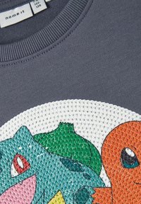 Sweat-shirt gris avec un graphique texturé représentant des personnages de dessin animé colorés : vert, bleu et orange. Encolure côtelée et détails de conception vibrants.
