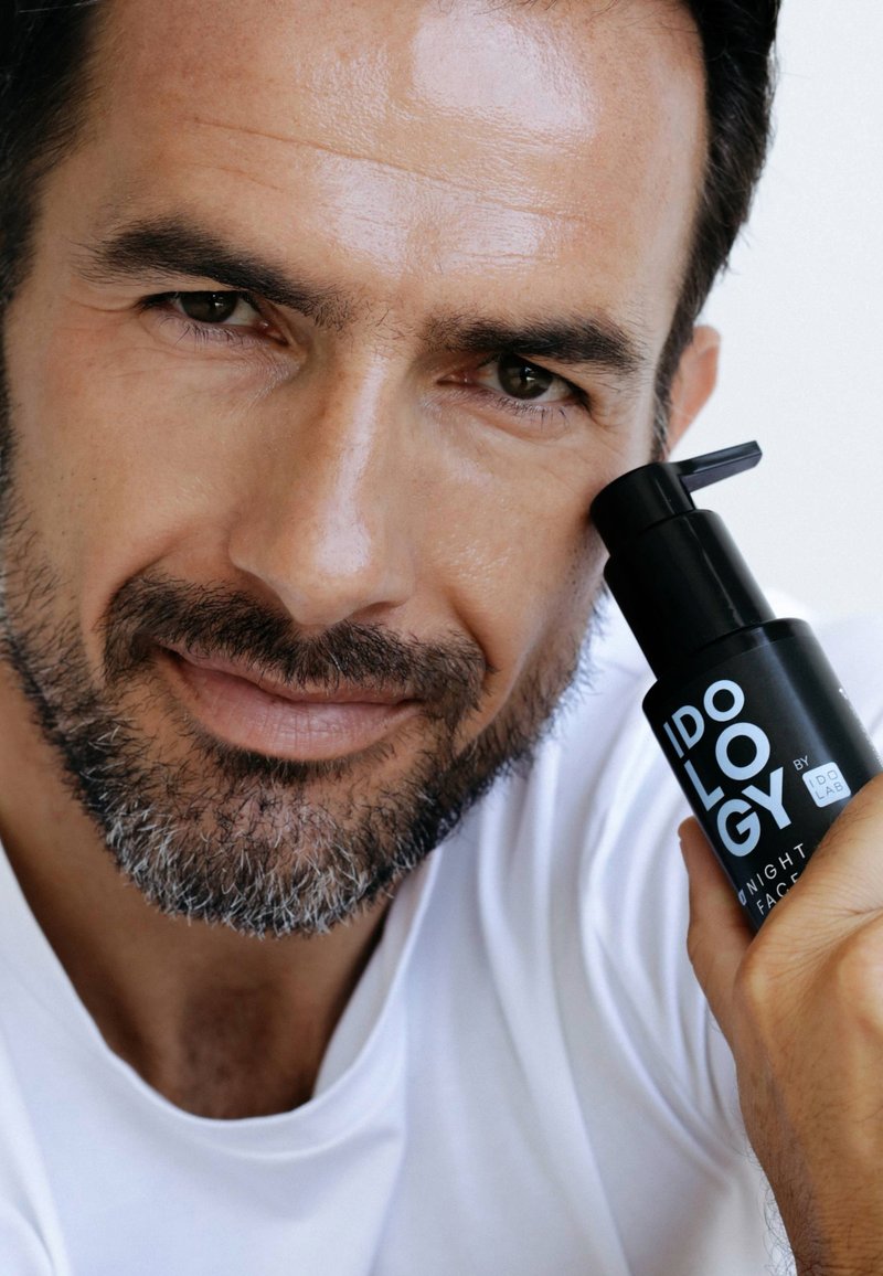 Czarna butelka do pielęgnacji skóry z pompką, oznaczona jako „Idology by IDO LAB”, trzymana obok twarzy mężczyzny z krótką brodą i gładką skórą.