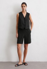 Schwarze Leinen-Weste ohne Ärmel mit V-Ausschnitt und Knöpfen, kombiniert mit passenden knielangen Shorts mit Fronttaschen und einem seitlichen Schlitz.