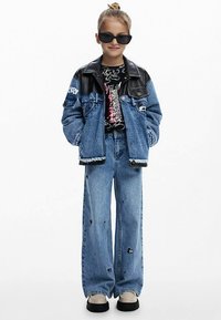 Jachetă din denim cu guler negru din piele, tricou negru cu imprimeu grafic, blugi largi cu aspect uzat și adidași groși. Ochelarii de soare completează ținuta.