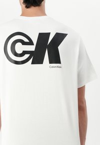 Balta medvilninė marškinėlių su dideliu juodu "CK" logotipu ir "Calvin Klein" tekstu nugaroje. Trumpomis rankovėmis, apvaliu kaklu.