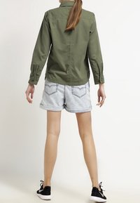 Camisa verde oliva de manga larga con detalles de botones, combinada con pantalones cortos de denim azul claro. Zapatillas negras completan el conjunto.