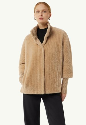 Giacca fuzzy beige con colletto sciallato, maniche corte e polsini a coste. Presenta una vestibilità e una consistenza morbide, abbinata a pantaloni neri.