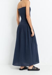 Robe maxi bustier bleu marine, avec un corsage smocké et une jupe plissée fluide. Associée à des sandales plates noires.
