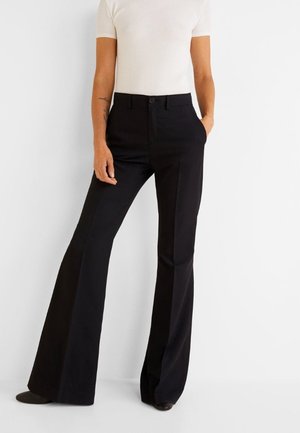 Personne portant un pantalon noir à jambes larges et un haut blanc ajusté à manches courtes, debout avec une main dans la poche devant un fond uni.