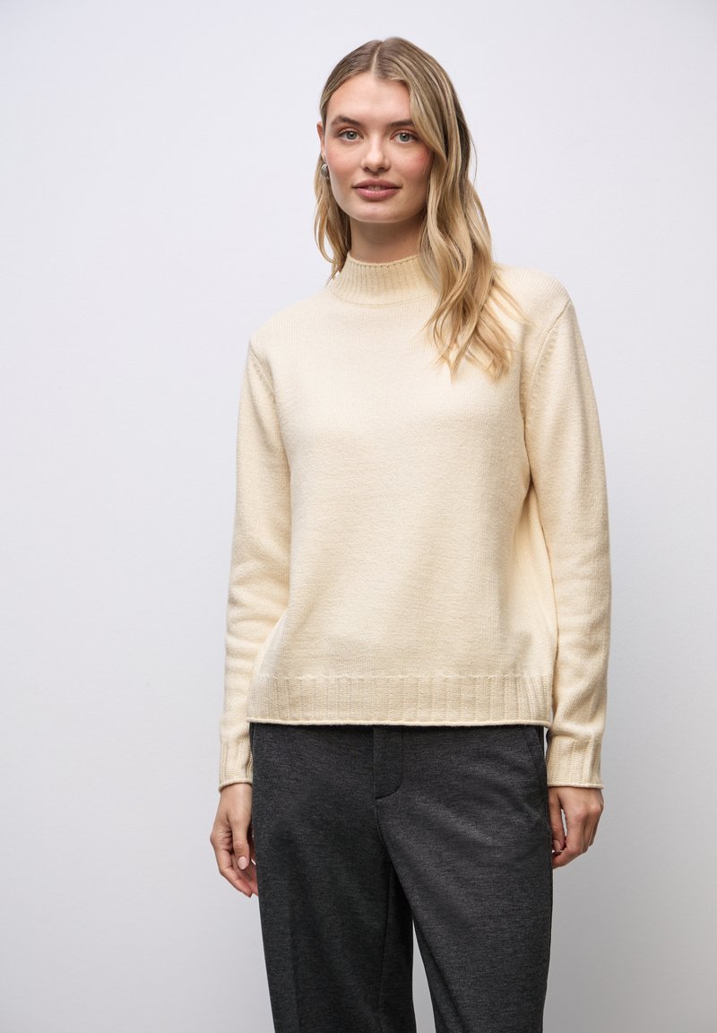 Pull en tricot de couleur crème avec un col montant et des poignets et un ourlet côtelés, associé à un pantalon sombre texturé.