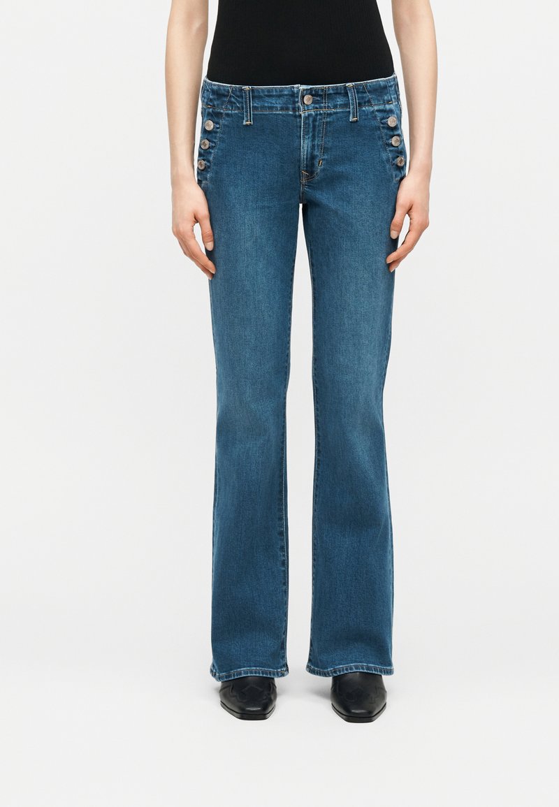 Μπλε τζιν σε γραμμή bootcut με έξι διακοσμητικά κουμπιά σε κάθε γοφό, φορεμένα με μαύρα παπούτσια με μυτερή μύτη και μαύρο τοπ.